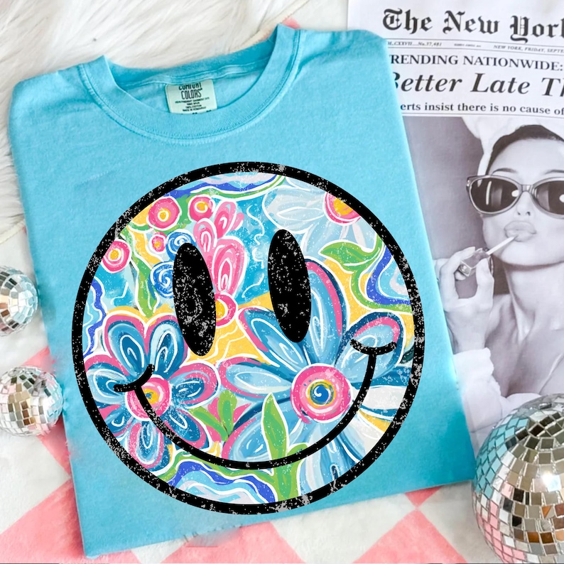 May include: A light blue t-shirt featuring a large smiley face design. The smiley face is filled with a floral pattern in shades of pink, blue, yellow, and green. The shirt is made of a soft material and is perfect for casual wear.