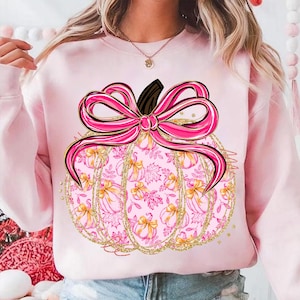 May include: Pink sweatshirt featuring a pumpkin design. The pumpkin is decorated with a pink bow, gold glitter accents, and a pattern of pumpkins and leaves. The design is centered on the front of the sweatshirt.