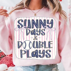 May include: Pink sweatshirt with the text "SUNNY DAYS & DOUBLE PLAYS" in navy blue, featuring a baseball graphic. The design includes bows and vertical pink stripes on a white background.