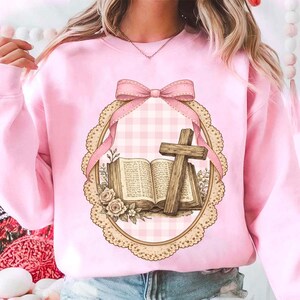 May include: A light pink sweatshirt featuring a framed graphic. The graphic includes an open book, a wooden cross, and floral accents. A pink bow and ribbon adorn the top of the frame, set against a pink and white gingham background.