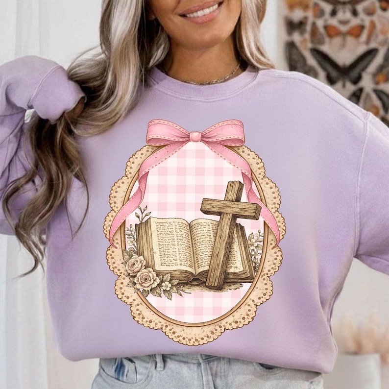 May include: A light purple sweatshirt featuring a decorative oval design with a pink bow, a cross, and an open book. The design has a pink gingham background and is framed by a scalloped edge with floral accents.