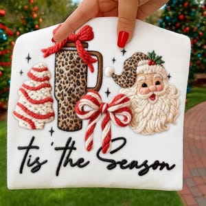 Puede incluir: Diseño navideño blanco con un vaso con estampado de leopardo, Papá Noel, un árbol de Navidad y un lazo rojo y blanco. La frase "Tis the Season" está escrita en letra negra. El diseño está sostenido por una mano con esmalte de uñas rojo.