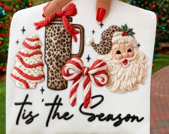 May include: White Christmas-themed design featuring a leopard print tumbler, Santa Claus, a Christmas tree, and a red and white bow. The phrase "Tis the Season" is written in black script. The design is held up by a hand with red nail polish.