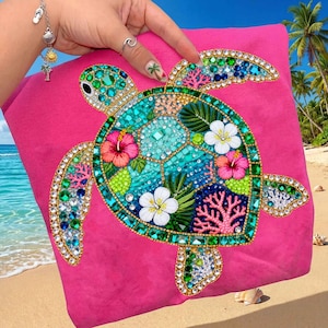 May include: A hot pink t-shirt featuring a detailed, bejeweled sea turtle design. The turtle's shell is a mosaic of turquoise, blue, and green gems, with accents of pink hibiscus flowers and coral. The design is outlined in gold.