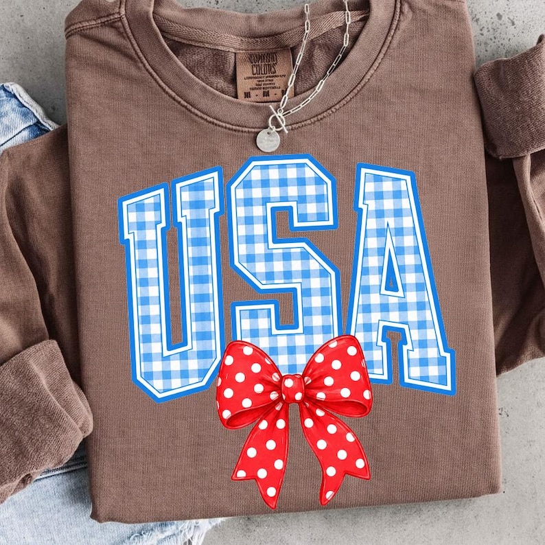 May include: A brown sweatshirt with the word "USA" in blue and white gingham print. A red bow with white polka dots is below the text. The sweatshirt has a crew neck and long sleeves.