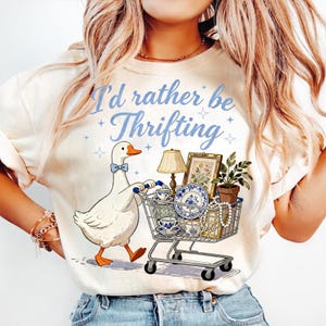 May include: Cream-colored t-shirt with the text "I'd rather be Thrifting" in blue script. A cartoon goose with a blue bow tie pushes a shopping cart filled with vintage items, including a lamp, framed art, and china.