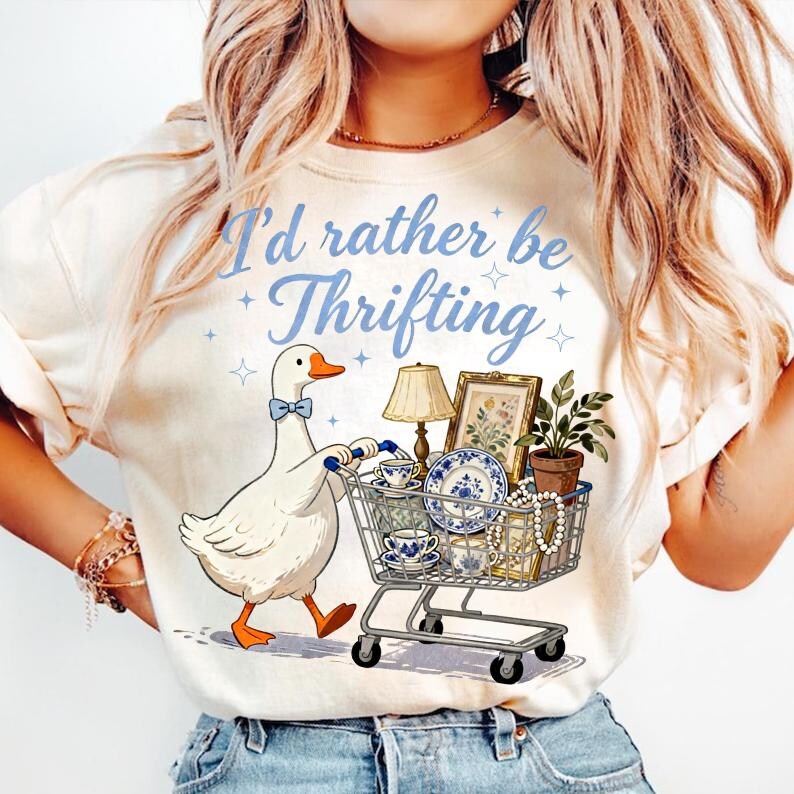 May include: Cream-colored t-shirt with the text "I'd rather be Thrifting" in blue script. A cartoon goose with a blue bow tie pushes a shopping cart filled with vintage items, including a lamp, framed art, and china.