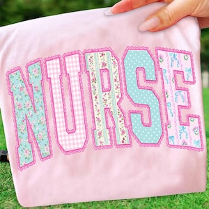 May include: A light pink t-shirt with the word "NURSE" in large, decorative letters. The letters are filled with floral, polka dot, and gingham patterns, outlined with pink lace trim. The shirt is held up by a hand.