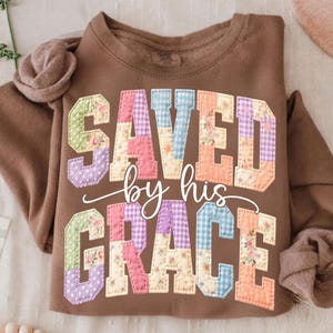 May include: A brown sweatshirt with the words "SAVED by his GRACE" in a patchwork design. The letters are in various colors and patterns, including floral and gingham prints. The sweatshirt has a crew neck and long sleeves.