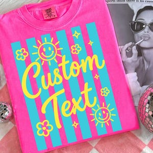 May include: Bright pink t-shirt with vertical turquoise stripes and yellow graphics. The design includes smiling suns, flowers, and stars, with the words "Custom Text" in a playful font. The shirt has a crew neck and a Comfort Colors tag.