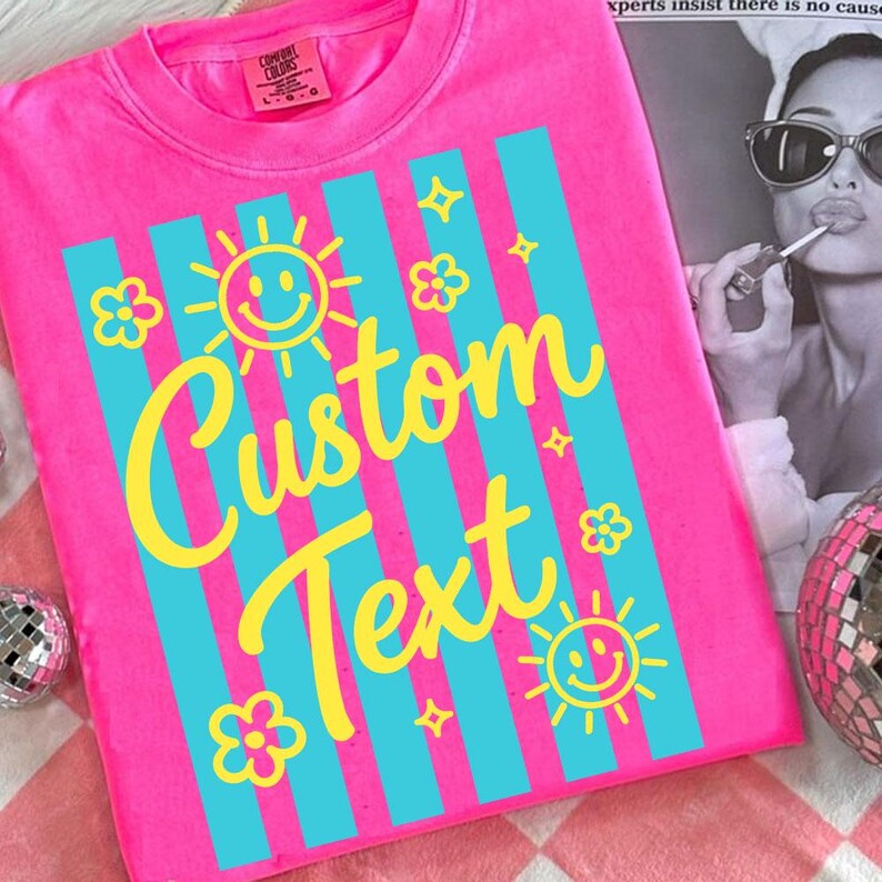 May include: Bright pink t-shirt with vertical turquoise stripes and yellow graphics. The design includes smiling suns, flowers, and stars, with the words "Custom Text" in a playful font. The shirt has a crew neck and a Comfort Colors tag.