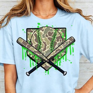 May include: Light blue t-shirt featuring a green and brown camouflage baseball and crossed bats graphic. The graphic has green paint splatters around it.