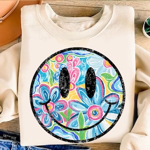 May include: Cream-colored sweatshirt featuring a large, distressed smiley face design. The smiley face is filled with a colorful floral pattern in shades of blue, pink, yellow, and green, outlined in black.