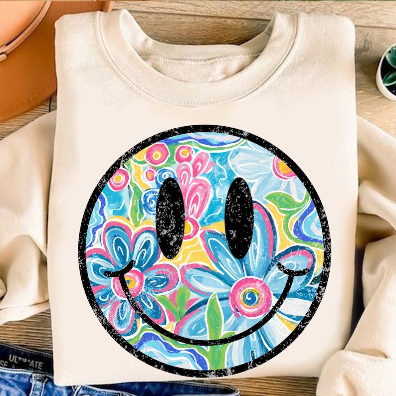 May include: Cream-colored sweatshirt featuring a large, distressed smiley face design. The smiley face is filled with a colorful floral pattern in shades of blue, pink, yellow, and green, outlined in black.