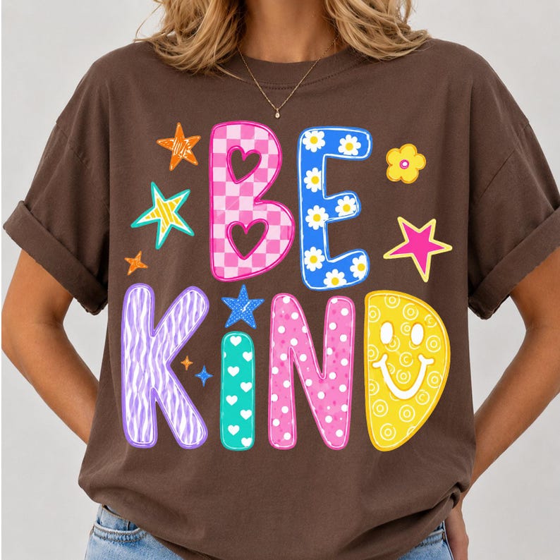 Op de afbeelding: Een donkerbruin t-shirt met de tekst BE KIND in grote, kleurrijke letters met patronen. De B is roze geblokt, de E blauw met madeliefjes, de K paars met zebrastrepen, de I turquoise met witte hartjes, de N roze met witte stippen en de D een gele smiley.