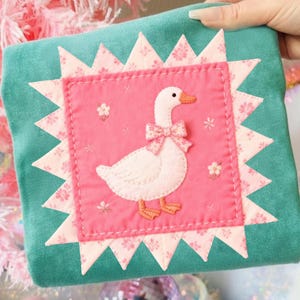 May include: A teal-colored, square-shaped pillow with a pink center featuring a white goose with a pink bow. The pink center is framed by a floral patterned border and a teal sawtooth edge. Small embroidered flowers are scattered around the goose.