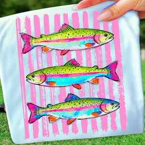 May include: A light blue t-shirt featuring three colorful rainbow trout illustrations. The fish have green, pink, blue, and orange accents. Vertical pink stripes are behind the fish.