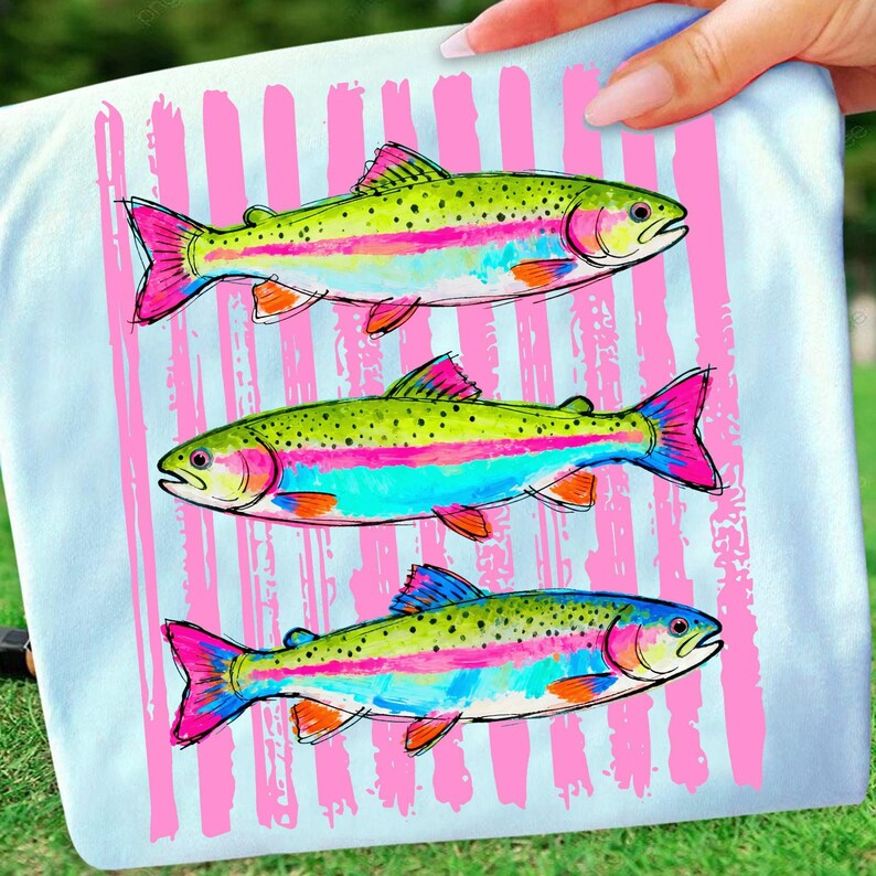 May include: A light blue t-shirt featuring three colorful rainbow trout illustrations. The fish have green, pink, blue, and orange accents. Vertical pink stripes are behind the fish.