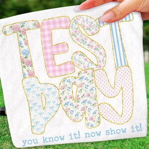 May include: White fabric with the words "TEST DAY" in large, colorful letters. The letters are decorated with floral, gingham, polka dot, and striped patterns, outlined in gold glitter. The text "you know it! now show it!" is printed below.