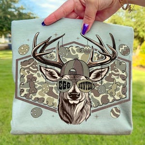 May include: A light blue t-shirt featuring a deer head wearing sunglasses with the text "EGG HUNTER". The deer is wearing a hat and has antlers. The background has a camouflage pattern and Easter egg designs.