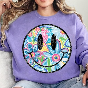 May include: A lilac-colored sweatshirt featuring a large smiley face design. The smiley face is a circle with a black outline and a floral pattern in the center, with blue, pink, and yellow flowers. The eyes are black ovals, and the mouth is a curved black line.