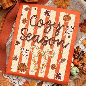 May include: An orange t-shirt featuring vertical white stripes with polka dots and the brown text Cozy Season. The design includes illustrations of pumpkins, autumn leaves, berries, and a decorative bow with a fall-themed pattern.