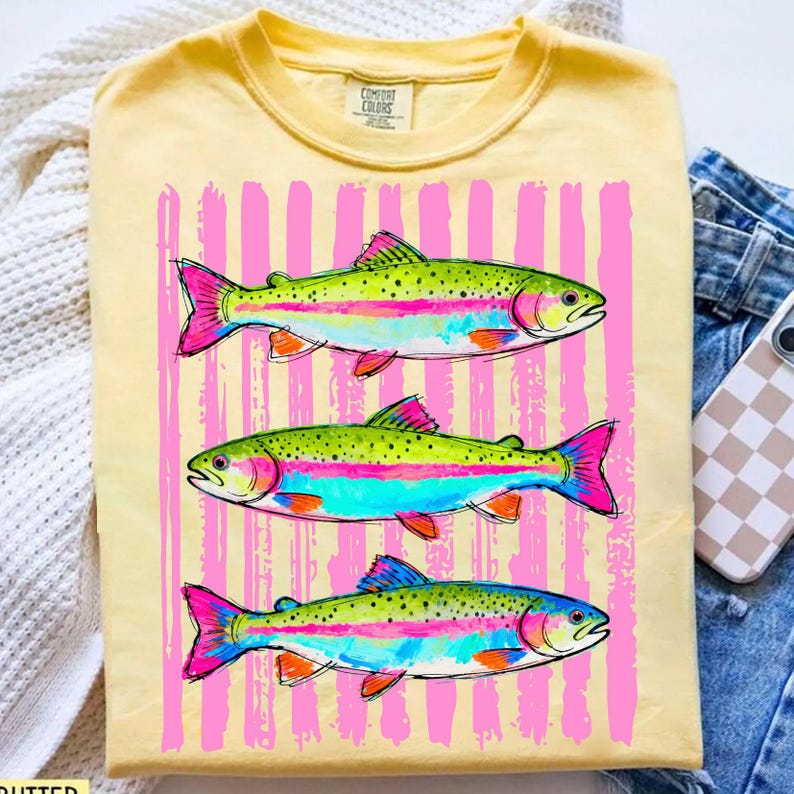 May include: Yellow t-shirt featuring a graphic of three rainbow trout with pink and blue accents, set against a backdrop of pink vertical stripes. The shirt is made of a soft material and has a crew neck.