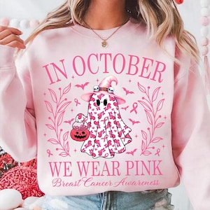 May include: Pink sweatshirt with the text "IN OCTOBER WE WEAR PINK Breast Cancer Awareness". The design features a ghost wearing a witch hat and holding a pumpkin basket, surrounded by bats and pink ribbons.