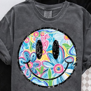 May include: Dark grey sweatshirt featuring a large smiley face with a floral design. The smiley face has black eyes and a black outline. The floral design includes blue, pink, yellow, and white flowers. The sweatshirt has a crew neck.