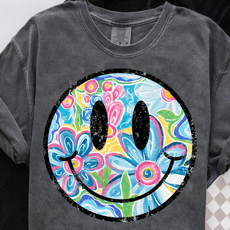 May include: Dark grey sweatshirt featuring a large smiley face with a floral design. The smiley face has black eyes and a black outline. The floral design includes blue, pink, yellow, and white flowers. The sweatshirt has a crew neck.