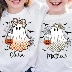 Puede incluir: Camisetas blancas con dise&ntilde;os de fantasmas de Halloween. Un fantasma tiene un lazo y un patr&oacute;n a cuadros naranja, el otro un sombrero de camuflaje y un patr&oacute;n a cuadros. Ambos sostienen bolsas de golosinas de calabaza. Murci&eacute;lagos negros y telara&ntilde;as rodean a los fantasmas. Los nombres Olivia y Matthew est&aacute;n impresos debajo.