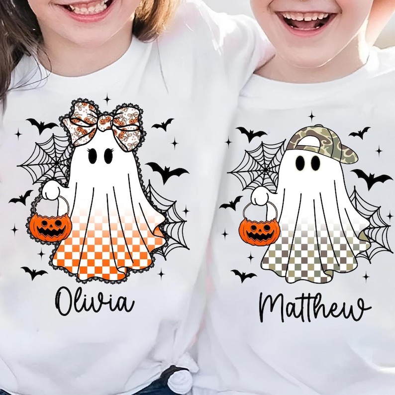 Puede incluir: Camisetas blancas con dise&ntilde;os de fantasmas de Halloween. Un fantasma tiene un lazo y un patr&oacute;n a cuadros naranja, el otro un sombrero de camuflaje y un patr&oacute;n a cuadros. Ambos sostienen bolsas de golosinas de calabaza. Murci&eacute;lagos negros y telara&ntilde;as rodean a los fantasmas. Los nombres Olivia y Matthew est&aacute;n impresos debajo.