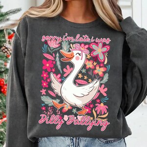 K&ouml;nnte beinhalten: Dunkelgraues Sweatshirt mit einem skurrilen Motiv einer wei&szlig;en Gans mit rosa Schleife, umgeben von rosa und floralen Elementen. Der Text "sorry i'm late i was Dilly Dallying" steht &uuml;ber und unter der Gans.