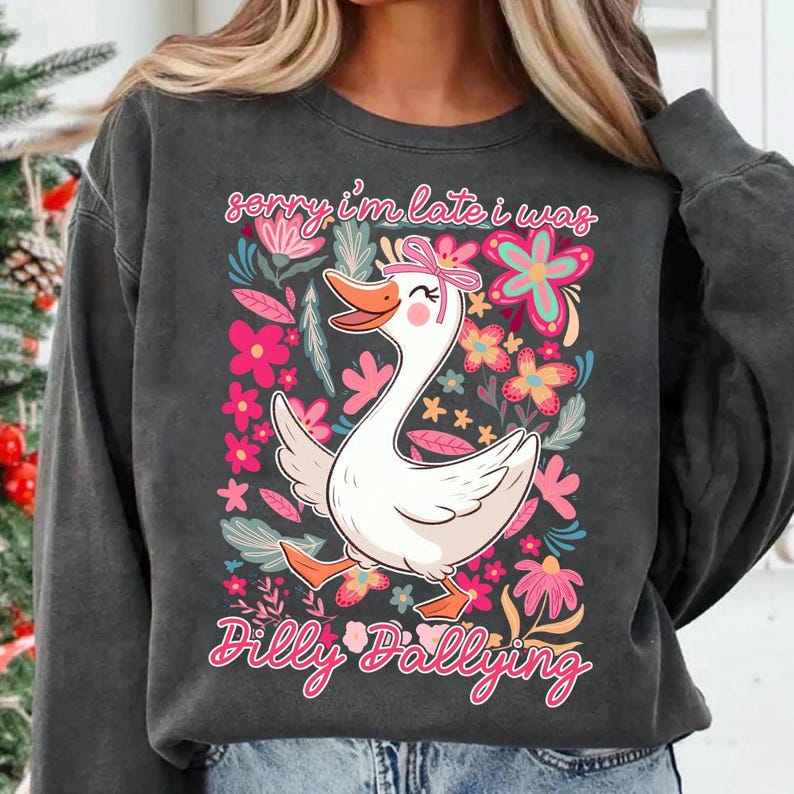 K&ouml;nnte beinhalten: Dunkelgraues Sweatshirt mit einem skurrilen Motiv einer wei&szlig;en Gans mit rosa Schleife, umgeben von rosa und floralen Elementen. Der Text "sorry i'm late i was Dilly Dallying" steht &uuml;ber und unter der Gans.