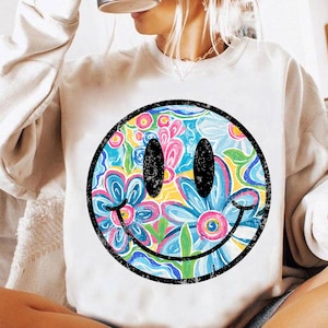 May include: A white sweatshirt featuring a large, colorful smiley face design. The smiley face has a black outline and is filled with a floral pattern in shades of blue, pink, yellow, and green. The sweatshirt is a casual, comfortable style.