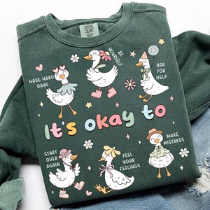 以下が含まれることがあります： 「It's okay to」や「Make Mistakes」などのフレーズとアクセサリーを身につけた漫画のアヒルのグラフィックデザインが施された濃い緑色のスウェットシャツ。デザインには花、星、「Have Hard Days」と「Be Yourself」の文字が含まれています。