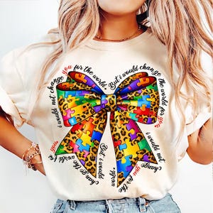 Puede incluir: Camiseta color crema con un gran diseño de lazo colorido. El lazo combina piezas de rompecabezas y estampados de leopardo. El texto rodea el lazo, que dice "No te cambiaría por el mundo."