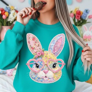May include: Teal sweatshirt featuring a colorful bunny face with heart-shaped glasses. The bunny design incorporates floral patterns and pink accents. The sweatshirt is a long-sleeved crew neck, perfect for Easter or spring.