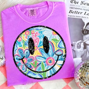 May include: A lilac Comfort Colors t-shirt featuring a large smiley face design. The smiley face has a black outline and is filled with a colorful floral pattern in shades of blue, pink, yellow, and green. The shirt is folded.