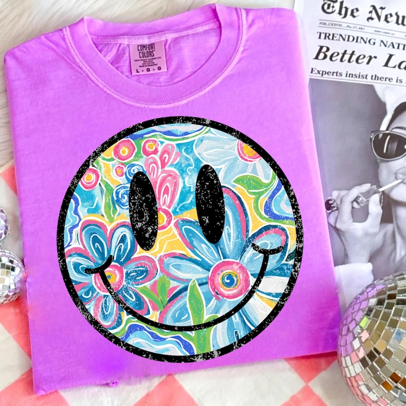 May include: A lilac Comfort Colors t-shirt featuring a large smiley face design. The smiley face has a black outline and is filled with a colorful floral pattern in shades of blue, pink, yellow, and green. The shirt is folded.
