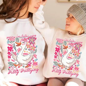 K&ouml;nnte beinhalten: Wei&szlig;e Sweatshirts mit einem bunten Aufdruck einer Gans, Blumen und dem Text "sorry i'm late i was Dilly Dallying". Die Gans hat eine rosa Schleife und einen orangefarbenen Schnabel. Die Sweatshirts sind wahrscheinlich f&uuml;r Eltern und Kind.