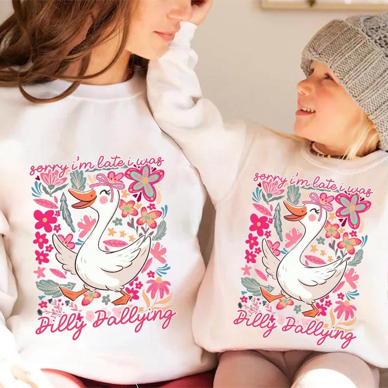 K&ouml;nnte beinhalten: Wei&szlig;e Sweatshirts mit einem bunten Aufdruck einer Gans, Blumen und dem Text "sorry i'm late i was Dilly Dallying". Die Gans hat eine rosa Schleife und einen orangefarbenen Schnabel. Die Sweatshirts sind wahrscheinlich f&uuml;r Eltern und Kind.