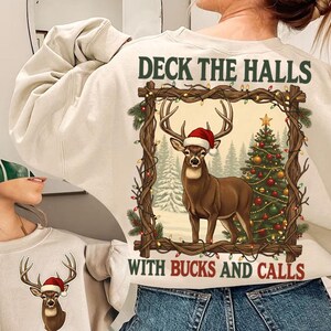 May include: Cream-colored sweatshirt with a Christmas-themed graphic. The graphic features a brown deer wearing a Santa hat, a decorated Christmas tree, and the text "DECK THE HALLS WITH BUCKS AND CALLS" in a rustic frame.