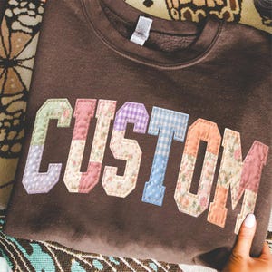 May include: A brown sweatshirt with the word "CUSTOM" appliqued across the front. The letters are made of various patterned fabrics, including floral, polka dot, and gingham designs. The sweatshirt is folded on a patterned surface.