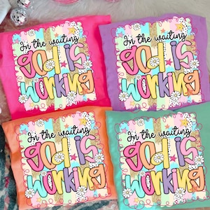 May include: Four colorful t-shirts with the text "In the waiting God is working" in a whimsical font. The shirts are in pink, purple, orange, and teal. Each shirt has a decorative border with stars and flowers.