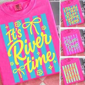 May include: Bright pink t-shirts with various river-themed designs. One shirt says "It's River Time" in yellow script with blue stripes and floral accents. Other shirts feature phrases like "Everybody on the River Getting Tipsy."