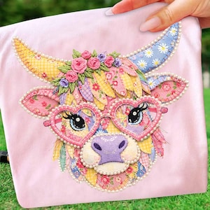 May include: A pink fabric item featuring a whimsical cow face design. The cow has heart-shaped glasses, floral accents, and a pearl-beaded trim. The design incorporates various patterned fabrics in shades of pink, yellow, and blue.