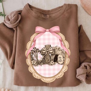 May include: A brown sweatshirt with an Easter graphic. The design features a cross, nails, and the text "He is Risen" inside a pink gingham oval frame, decorated with a pink bow and lace edging. The sweatshirt has a crew neck and long sleeves.