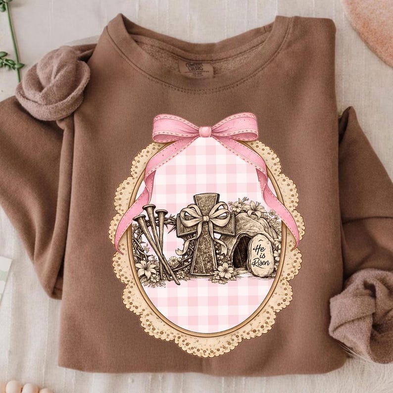May include: A brown sweatshirt with an Easter graphic. The design features a cross, nails, and the text "He is Risen" inside a pink gingham oval frame, decorated with a pink bow and lace edging. The sweatshirt has a crew neck and long sleeves.