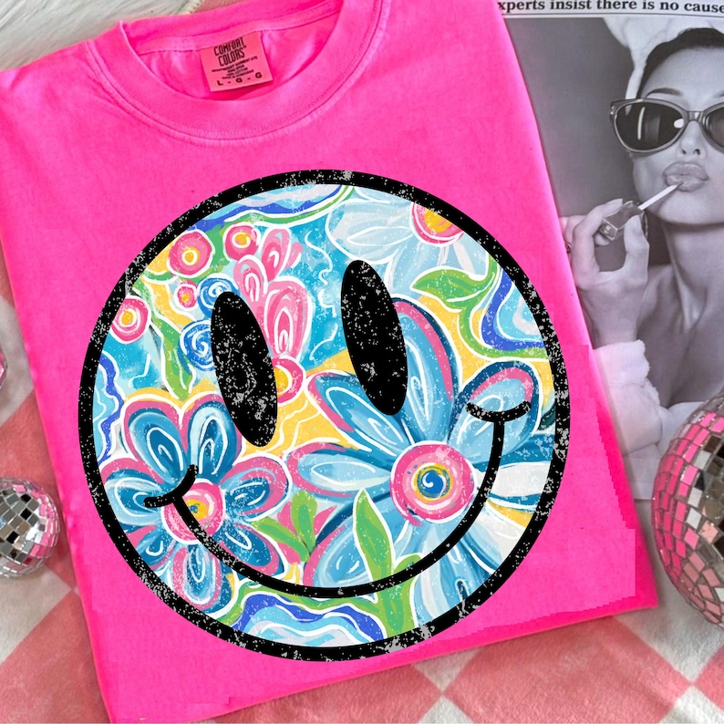 May include: Bright pink Comfort Colors t-shirt featuring a large smiley face graphic. The smiley face has black eyes and a black smile, with a floral design in the center. The floral design includes blue, pink, and yellow flowers.